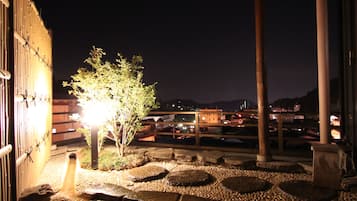 Un espacio al aire libre con un sendero de piedra que lleva a un pequeño estanque, rodeado de rocas y plantas, bajo una estructura de madera de noche.