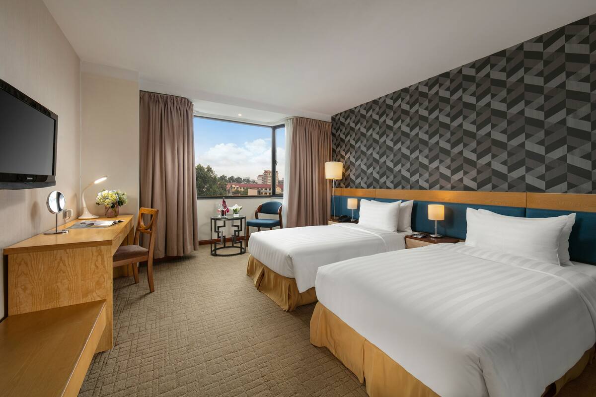 Una habitación de hotel con dos camas, un escritorio con una silla, un televisor y un ventanal con cortinas.