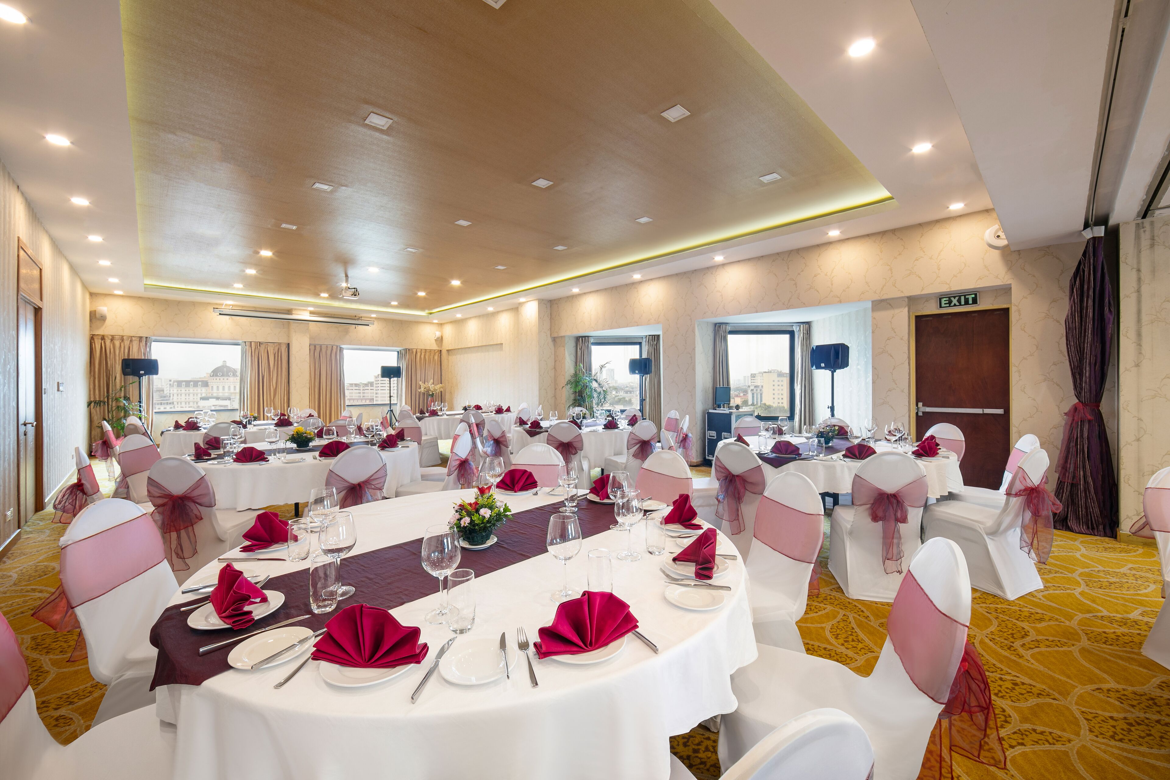 Un salón de banquetes con mesas redondas, sillas blancas adornadas con cintas rosas y suelo alfombrado.