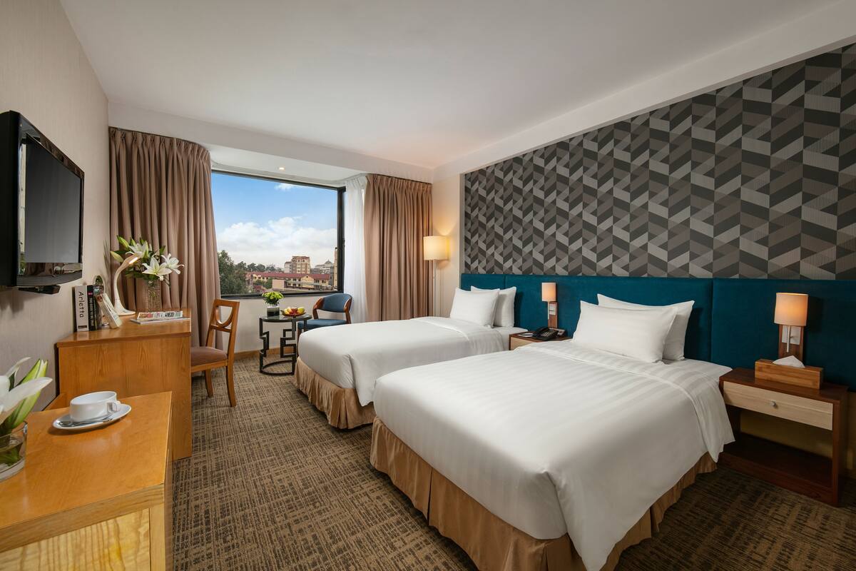 Habitación de hotel con dos camas, un escritorio con silla, televisor y vista a la ciudad.