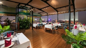Un restaurant avec des tables dressées pour le service, de grandes fenêtres donnant sur la ville et des plantes d’intérieur.
