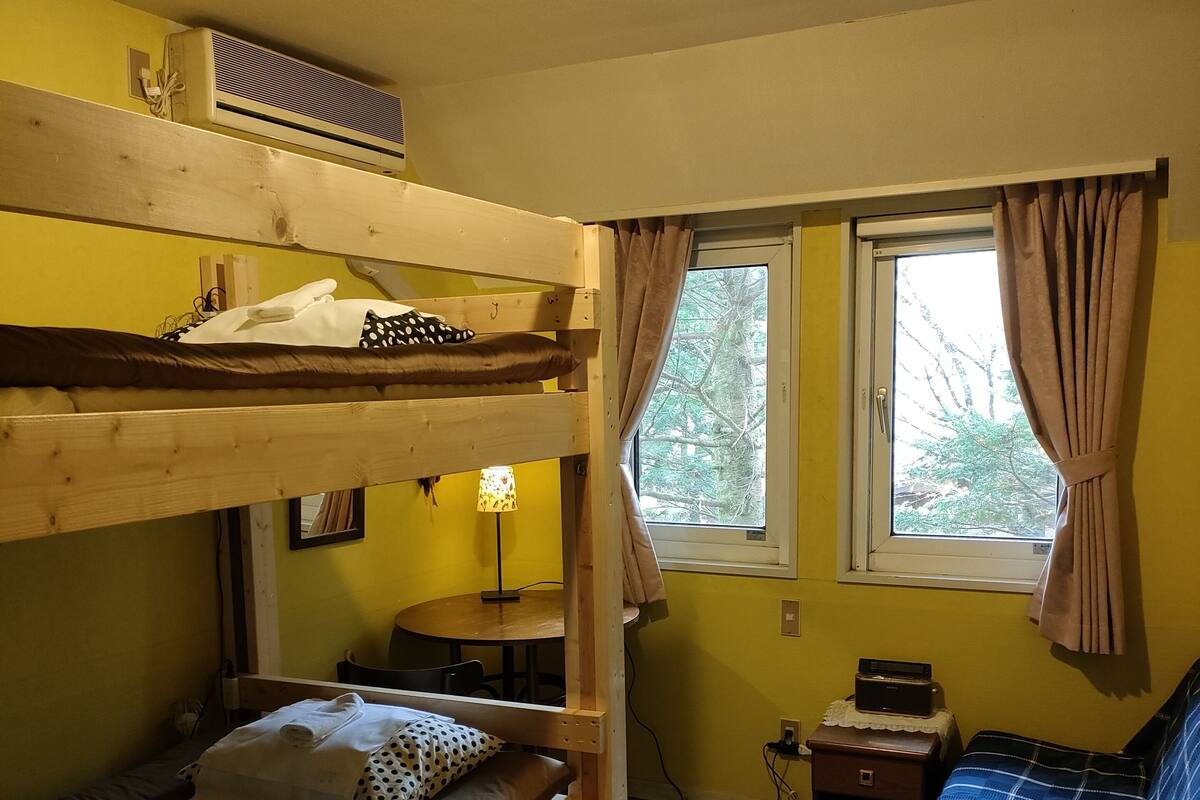 A dormitory room with bunk beds, a small table, a lamp, a window with curtains, and a radiator.