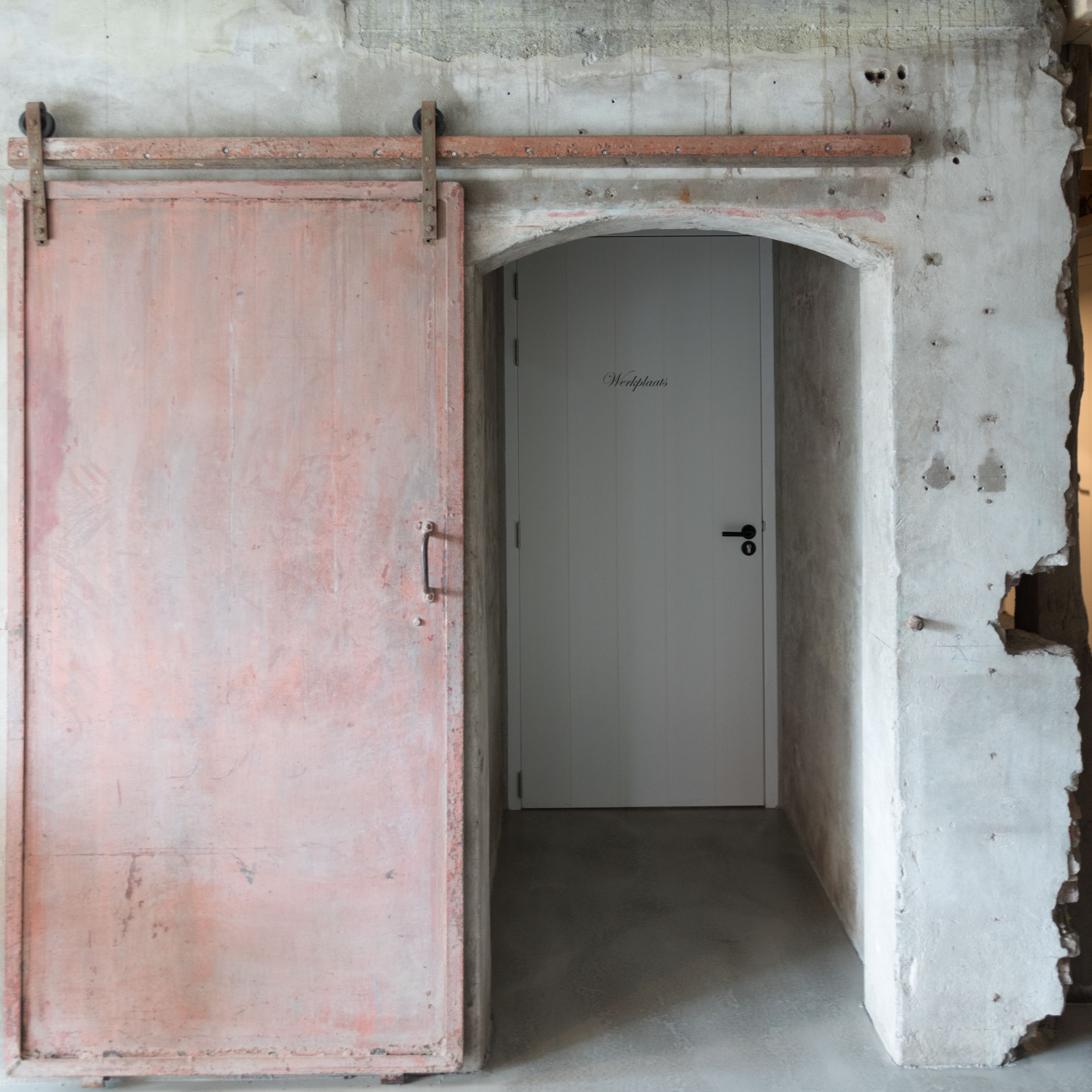 Una stanza con una porta rosa, una porta bianca e un muro in cemento con evidenti danni.