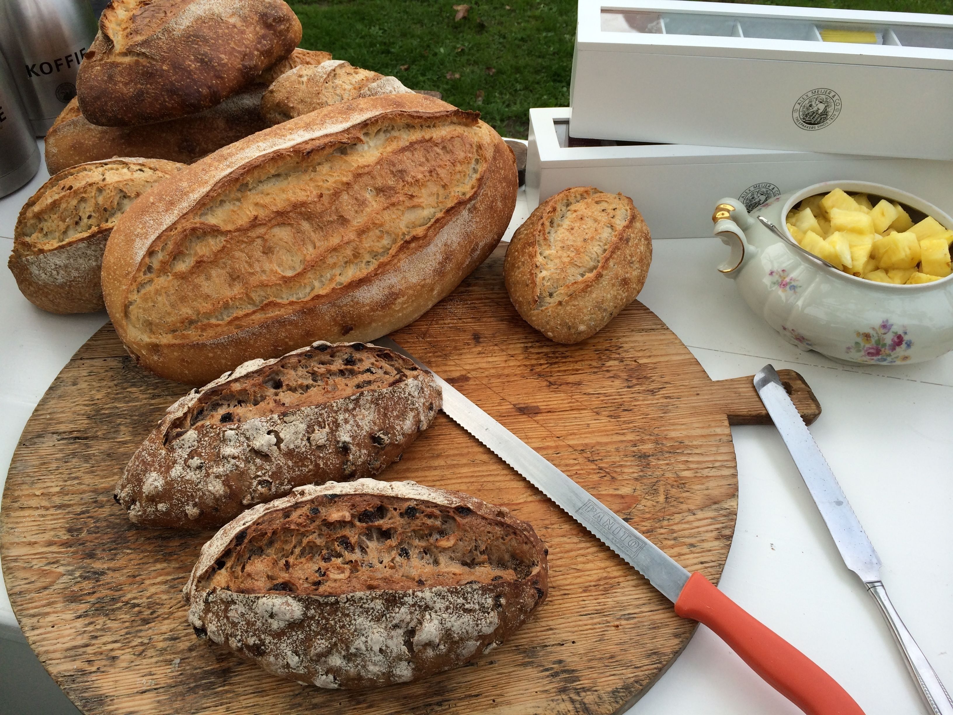 Una tabla de madera con varias hogazas de pan, un cuchillo y una tetera con trozos de piña.