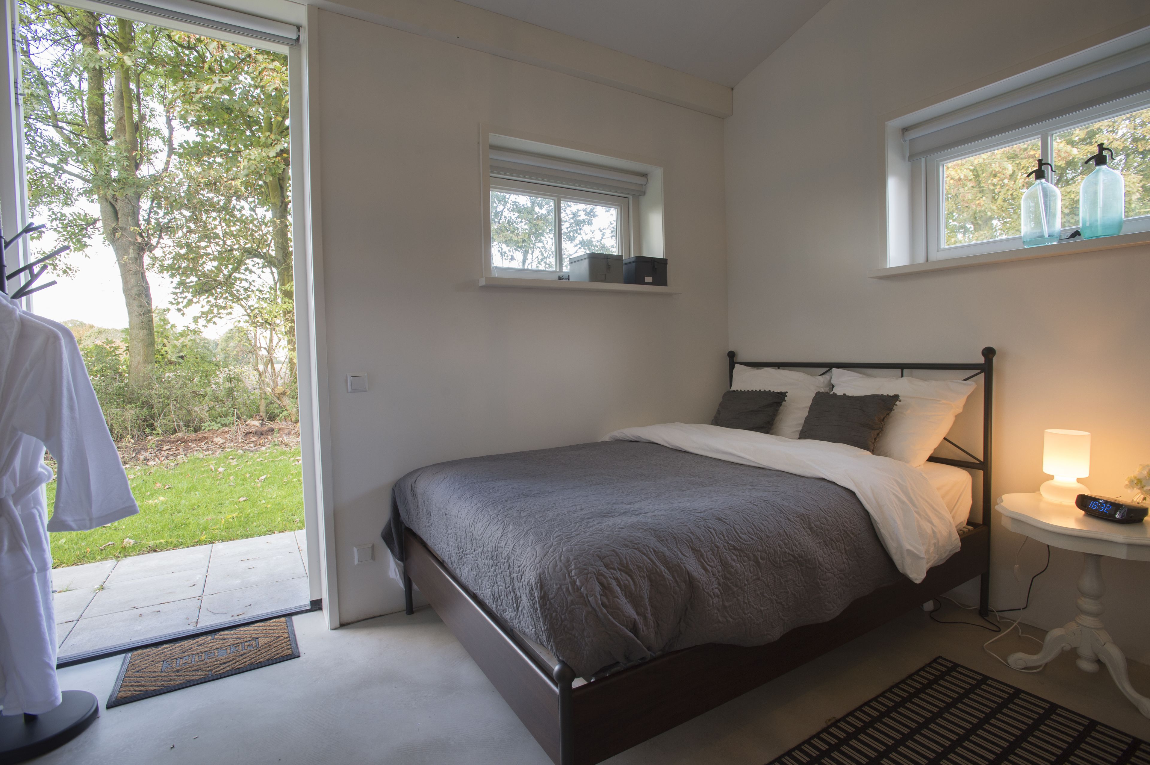 A bedroom with a bed, a bedside table with a lamp, a window with bottles, and a view of trees.
