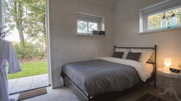 A bedroom with a bed, a bedside table with a lamp, a window with bottles, and a view of trees.