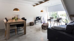 A modern living room with a wooden dining table, two chairs, a sofa, and a TV. There are wicker ottomans and pendant lights.