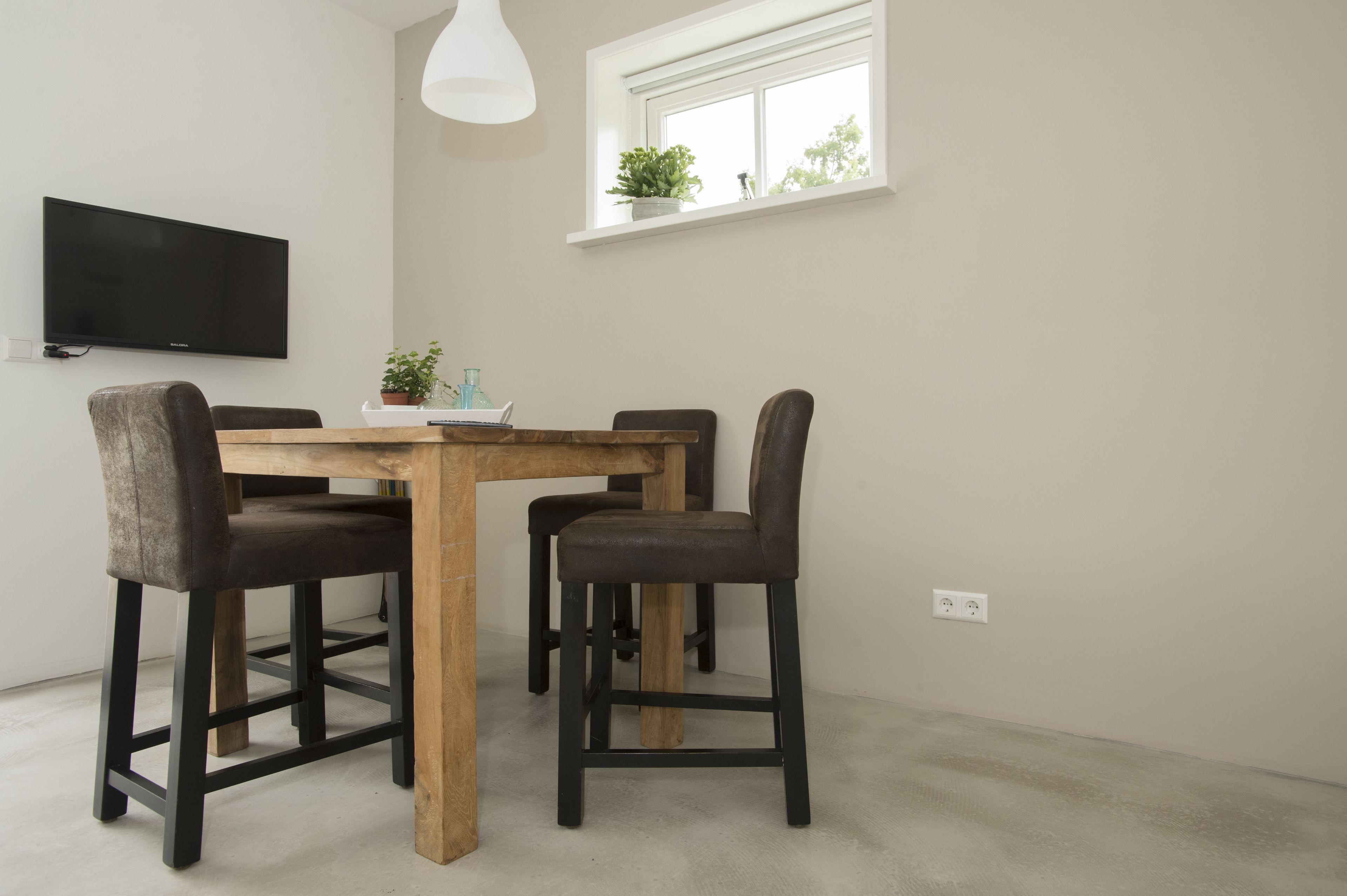 Un comedor con una mesa de madera y cuatro sillas, un televisor de pantalla plana montado en la pared, y una ventana con una planta en el alféizar.