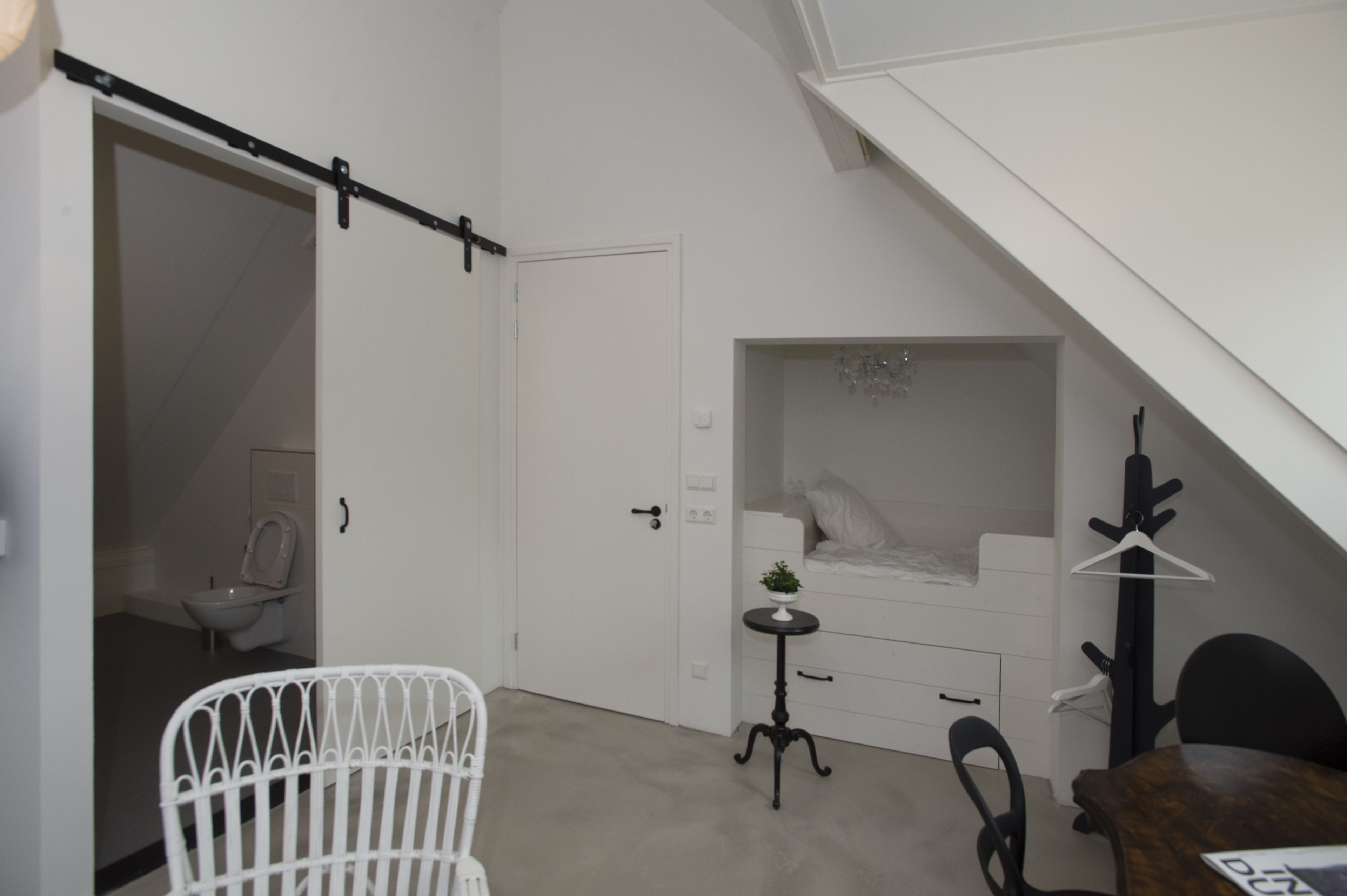 A small, white room with a bed, a small table with a plant, a chair, and a coat rack.
