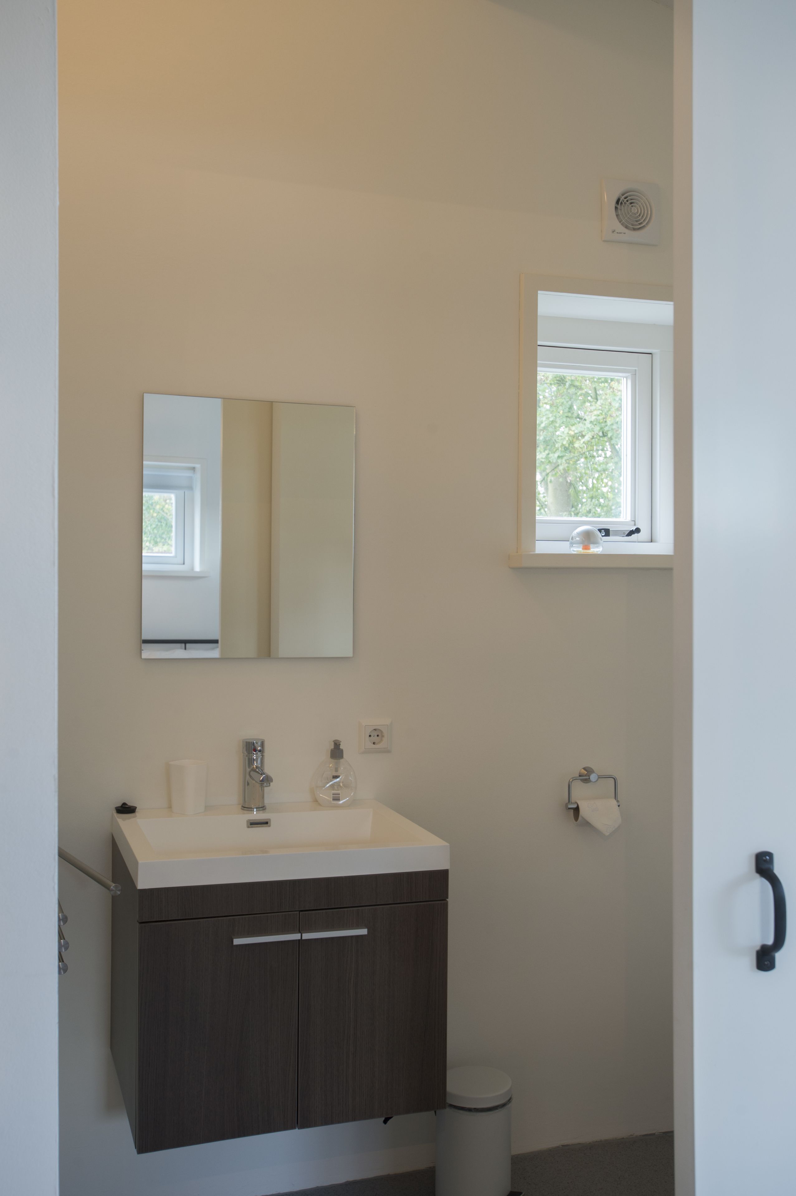 Een moderne badkamer met een witte wastafel, donkere kastjes, een spiegel, een raam en een toilet.