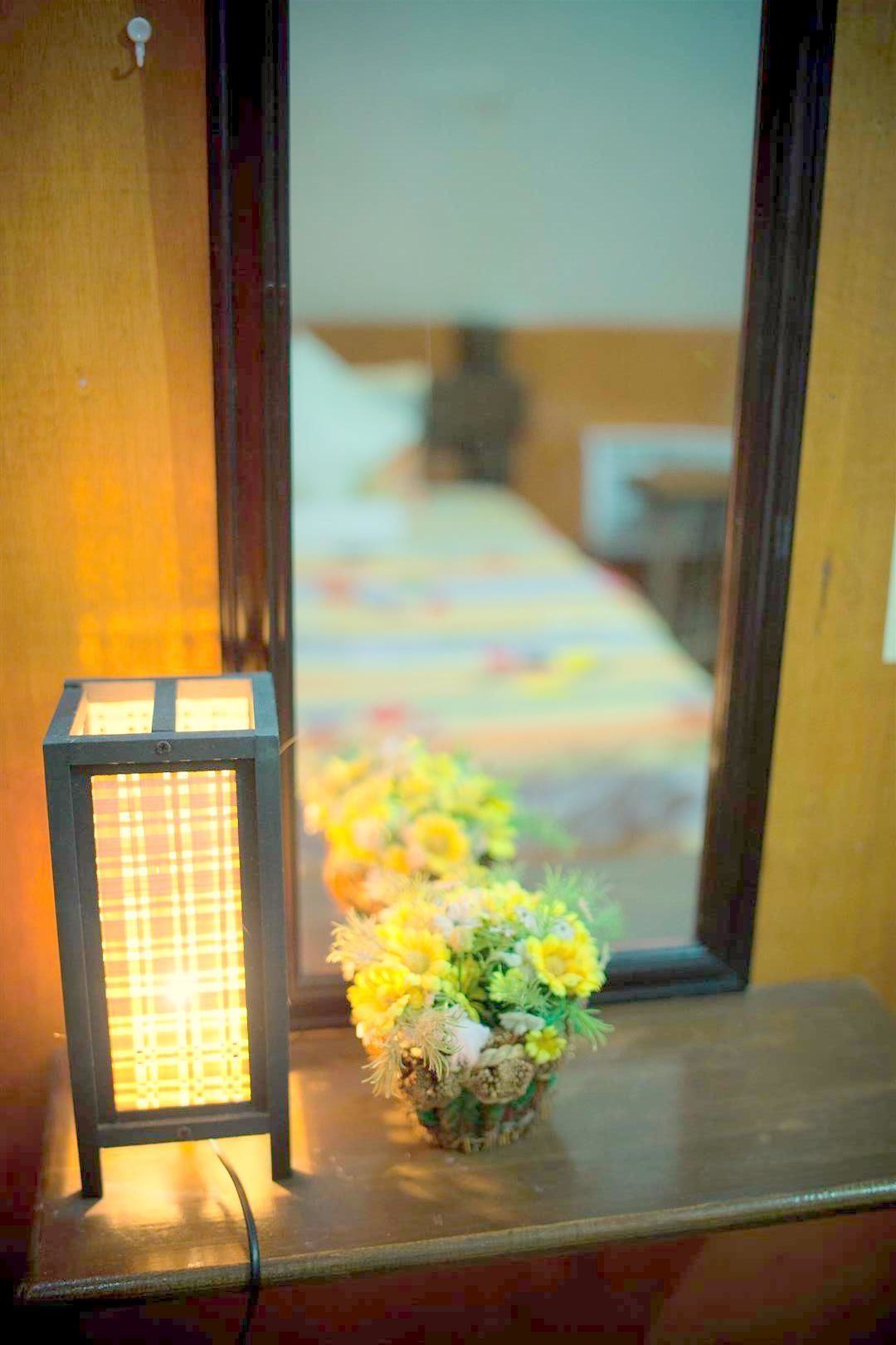 A window with a mirror reflecting a bed and a bedside table with a lamp and a flower arrangement.