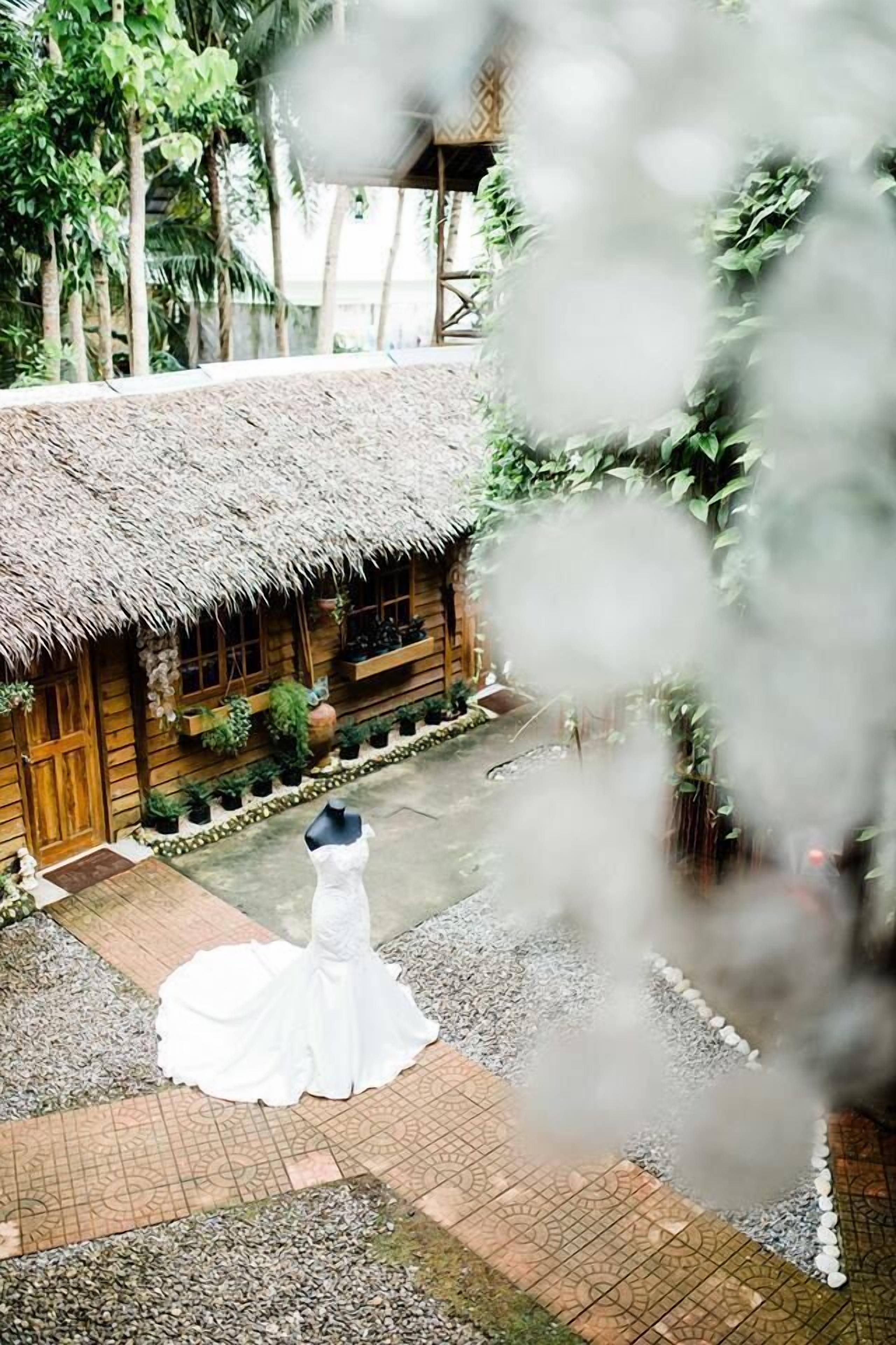 A rustic wooden building with a thatched roof, surrounded by greenery and potted plants.