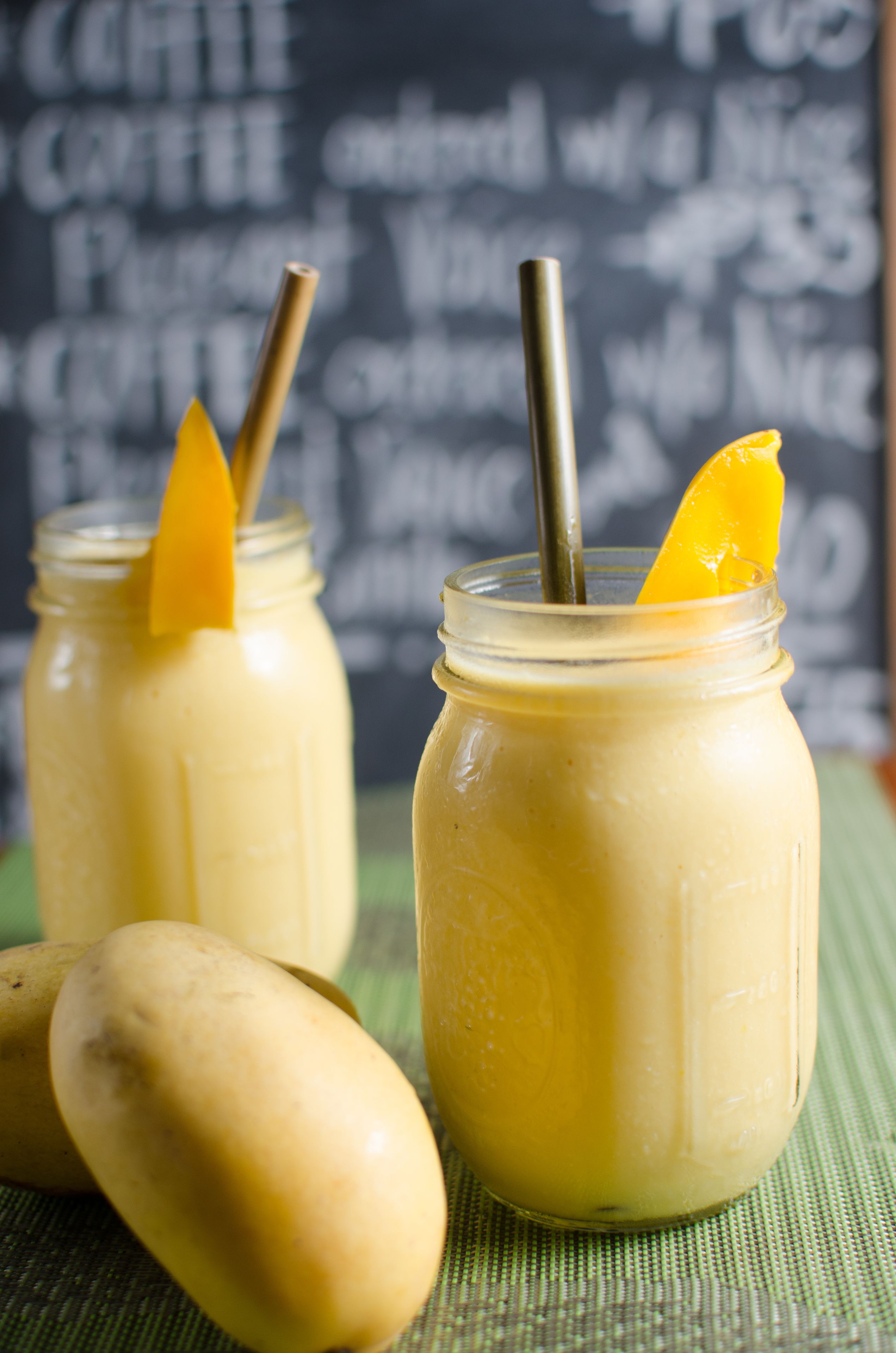 Zwei Gläser mit gelbem Smoothie, mit Orangenschalen und Trinkhalmen, auf einer grünen Matte vor einem Kreidetafelhintergrund.