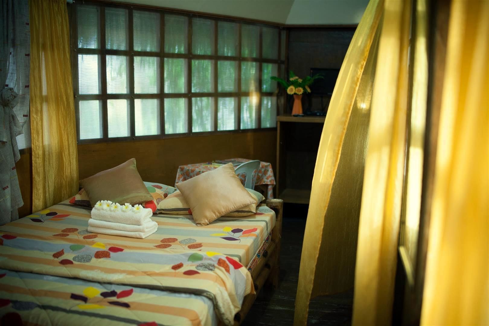 A hotel room with a large bed, multiple pillows, a bedside table with a vase of flowers, and a window with blinds.