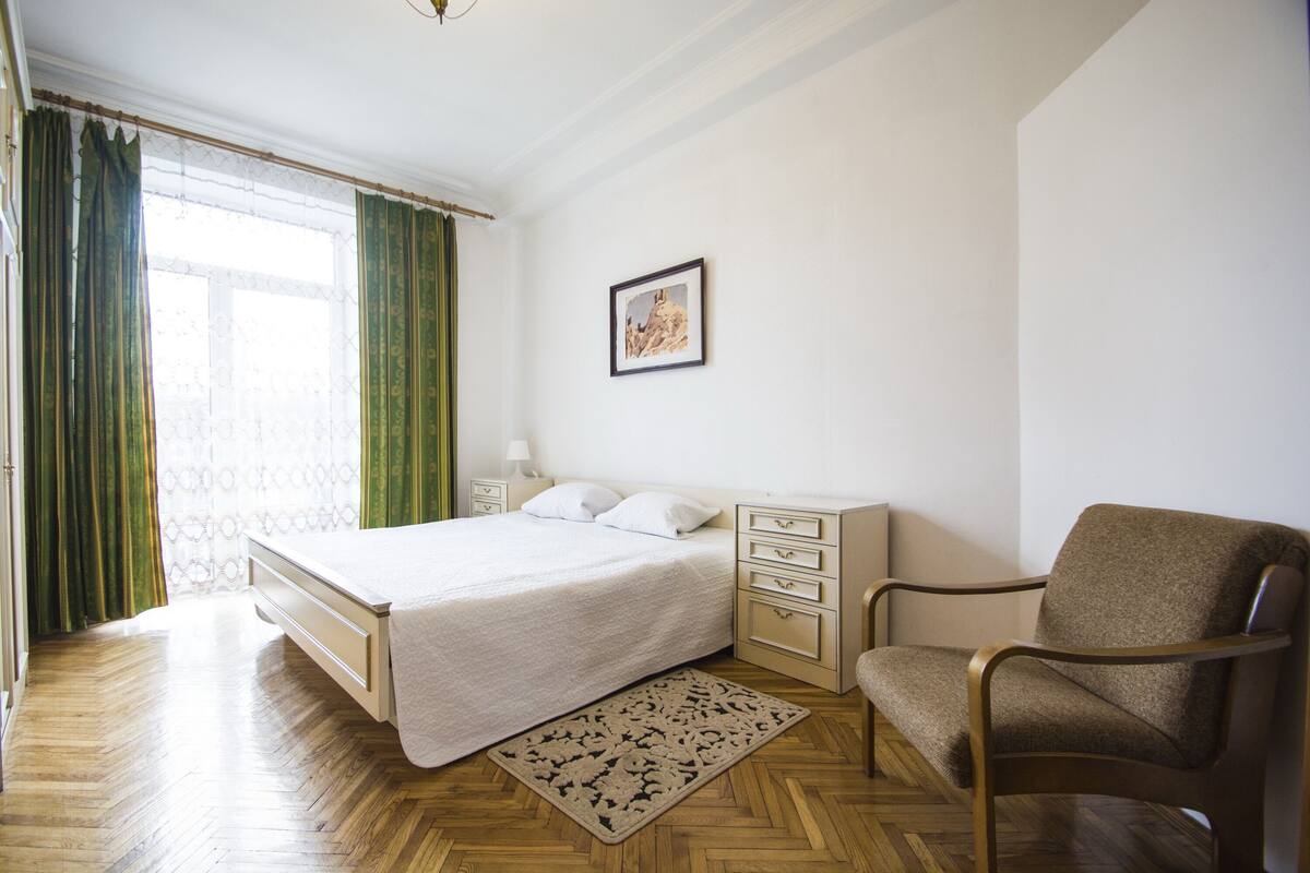 Un dormitorio con cama, una silla, una mesita de noche, una lámpara, un cuadro enmarcado en la pared y una ventana con cortinas.