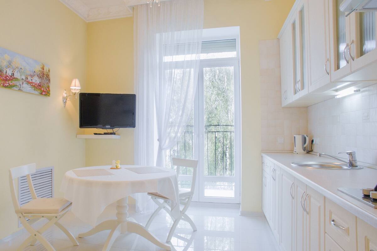 Una cocina luminosa con armarios blancos, una mesa de comedor con sillas, un televisor montado sobre la mesa y una ventana con cortinas traslúcidas.