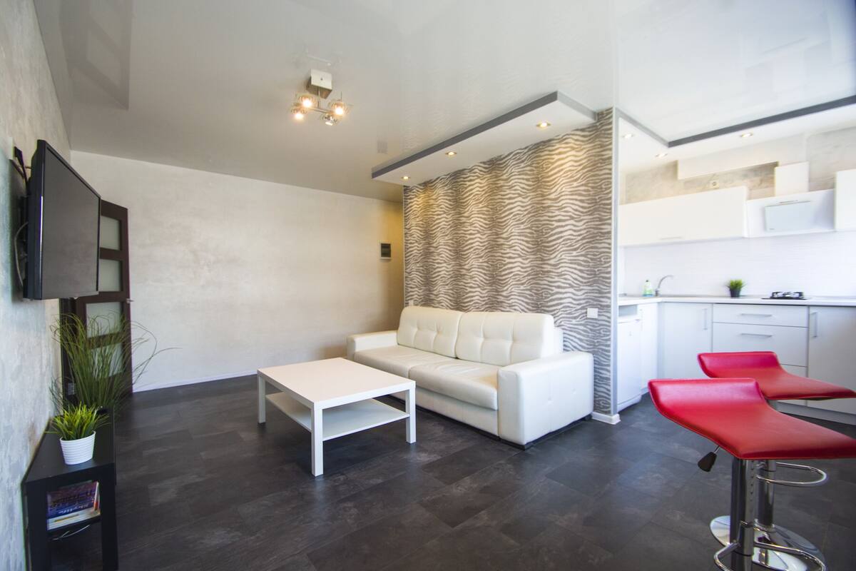 Una sala de estar moderna con un sofá blanco, una mesita pequeña blanca y taburetes rojos. Unas cortinas con estampado de cebra separan el espacio de la cocina.