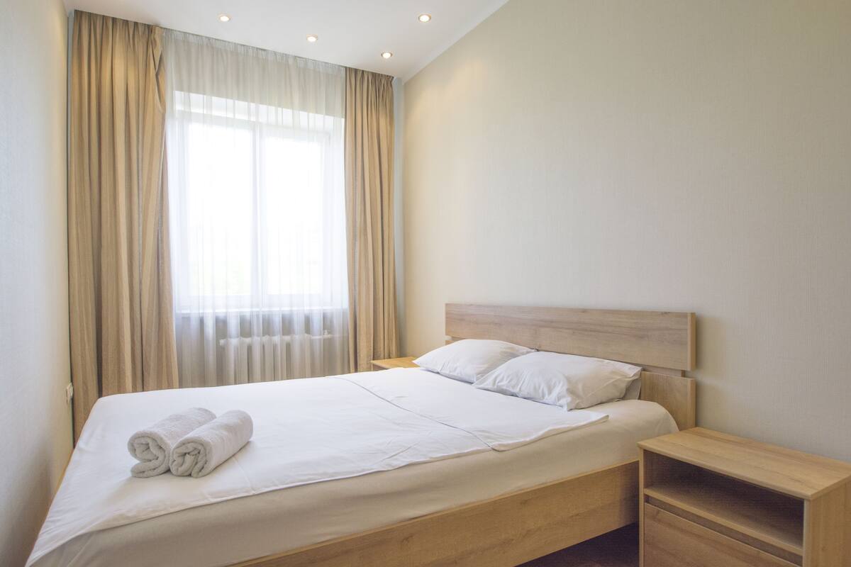 Una cama bien tendida con sábanas blancas y una toalla enrollada, una mesita de noche de madera con cajón, y una ventana con cortinas color beige.