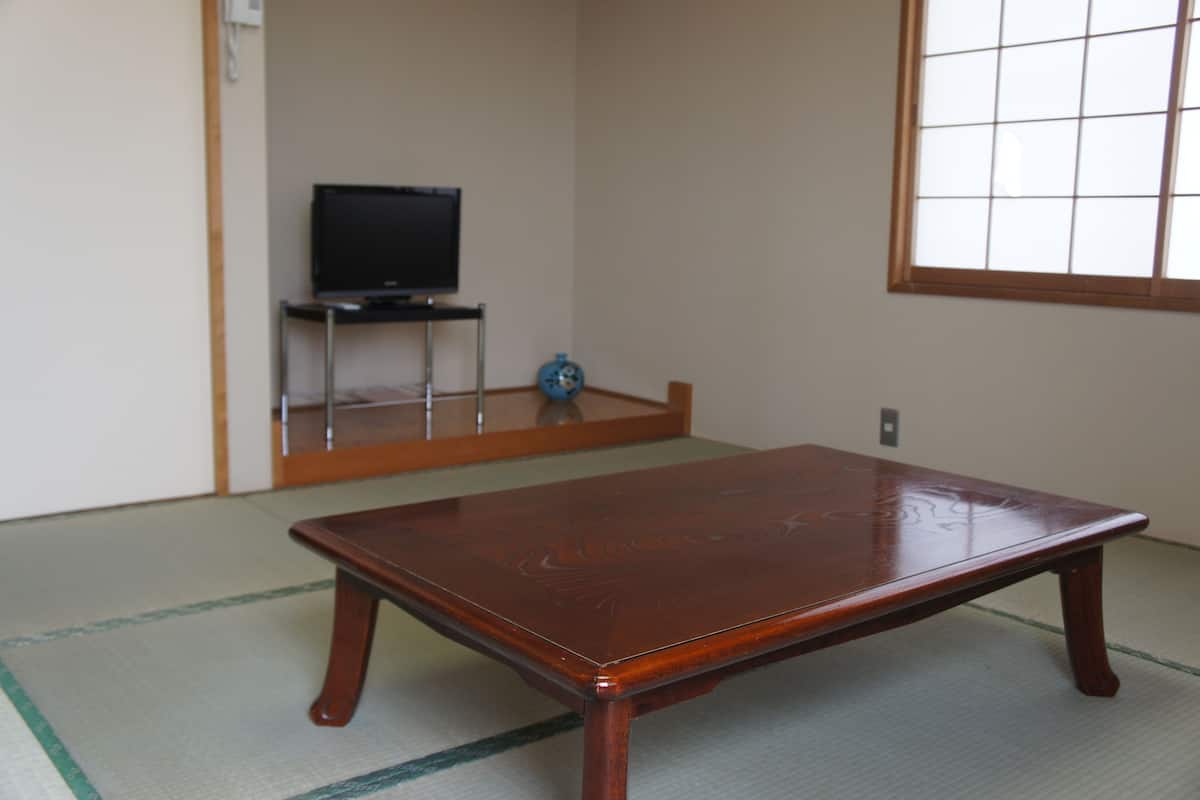 Een traditionele Japanse kamer met tatami vloeren, een houten tafel, een televisie op een standaard en een raam met een rasterpatroon.