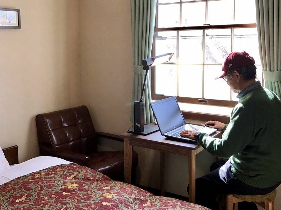 A hotel room with a bed, a desk with a laptop, a chair, and a window with curtains.