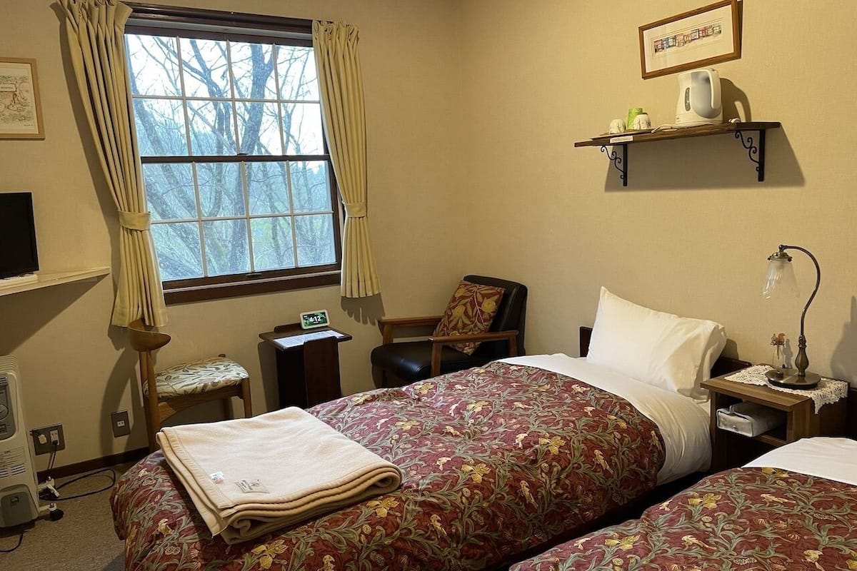 Een hotelkamer met twee bedden, een bureau, een stoel, een raam met gordijnen, een plank met een waterkoker en een nachtkastje met een lamp.