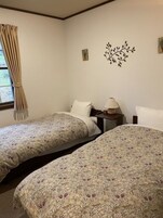 A room with two beds, a window with curtains, a bedside table with a lamp, and a wall decoration.