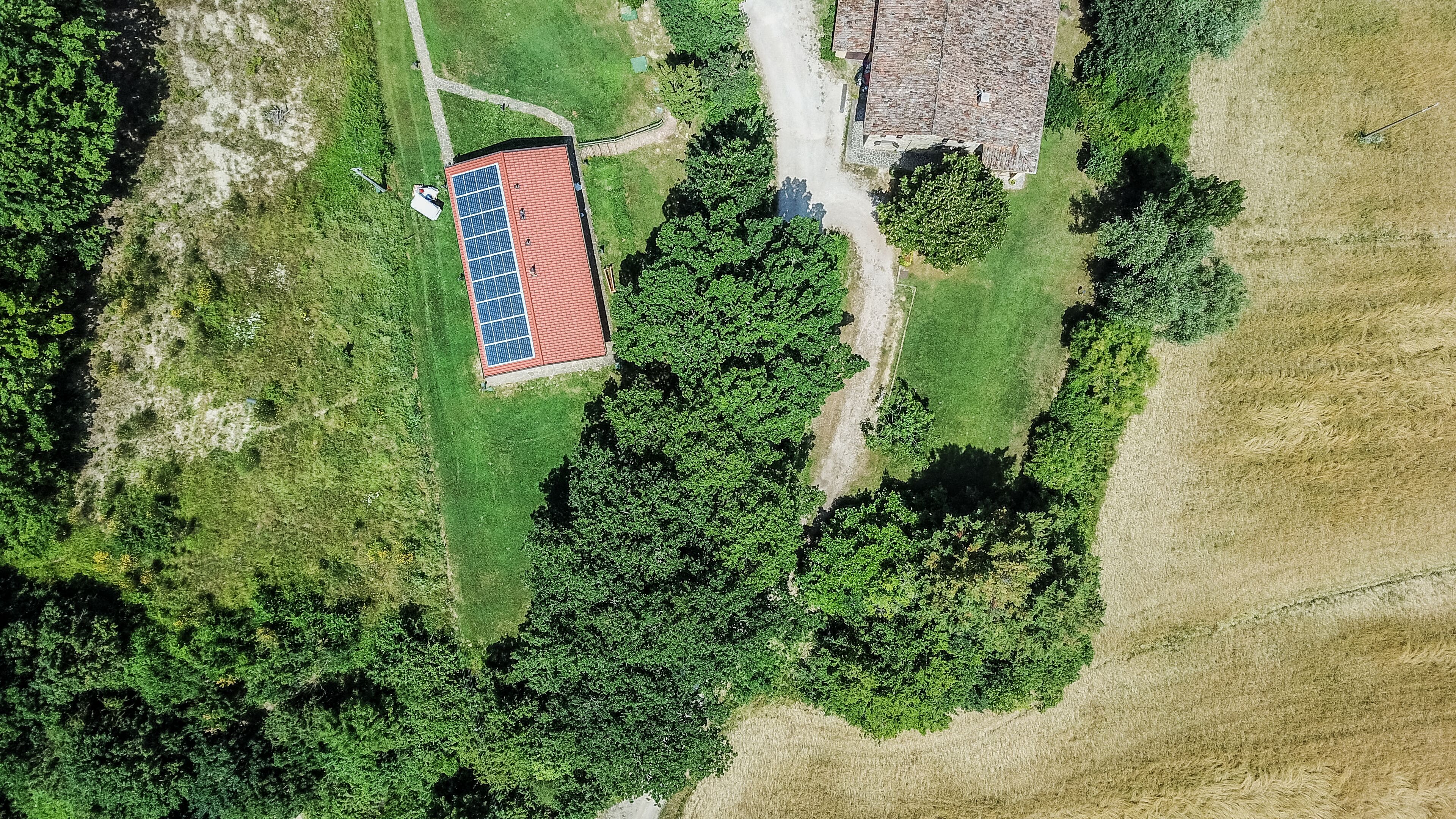 An aerial view of a property with a solar panel-equipped building, a dirt path, and surrounding greenery.