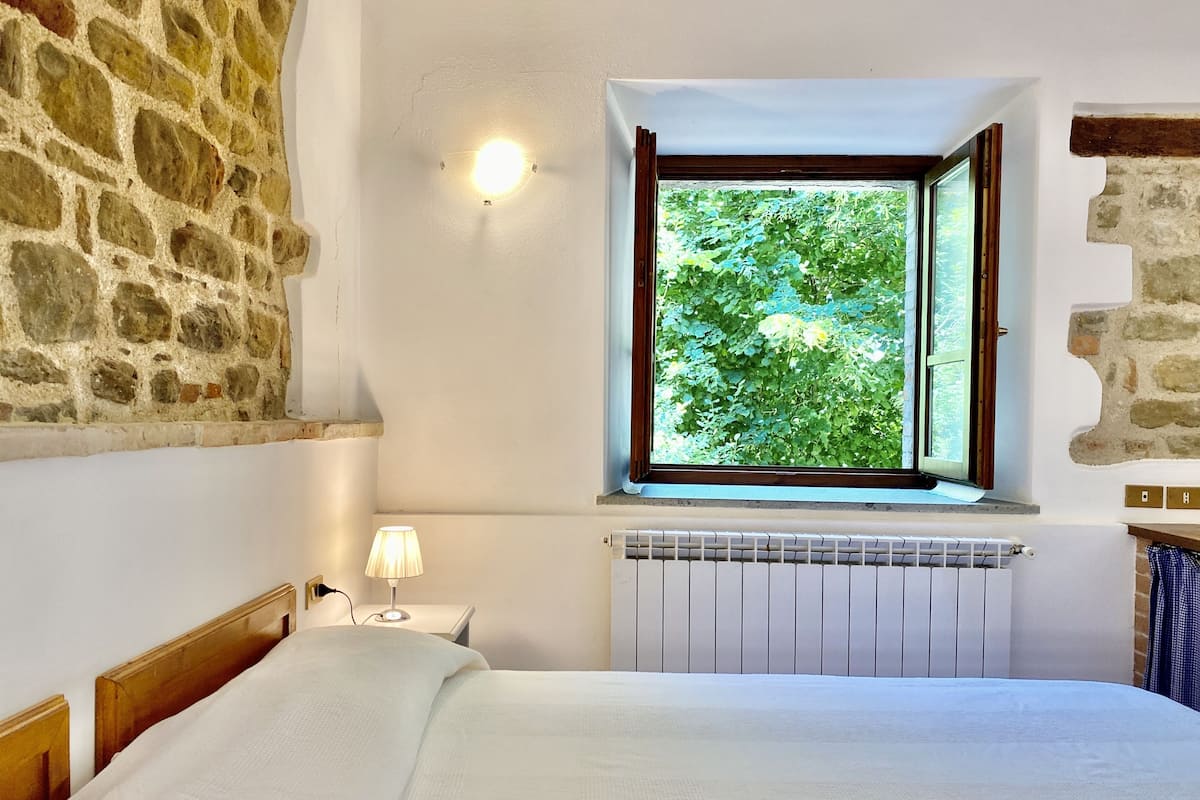A bedroom with a stone wall, a window with a view of greenery, a bedside lamp, and a radiator.
