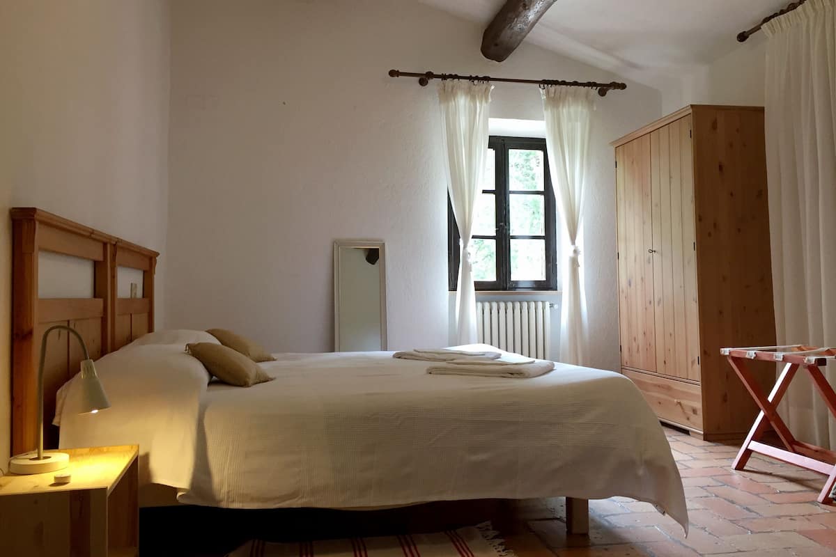 A bedroom with a bed, a nightstand, a mirror, a window with curtains, a radiator, and a wooden wardrobe.