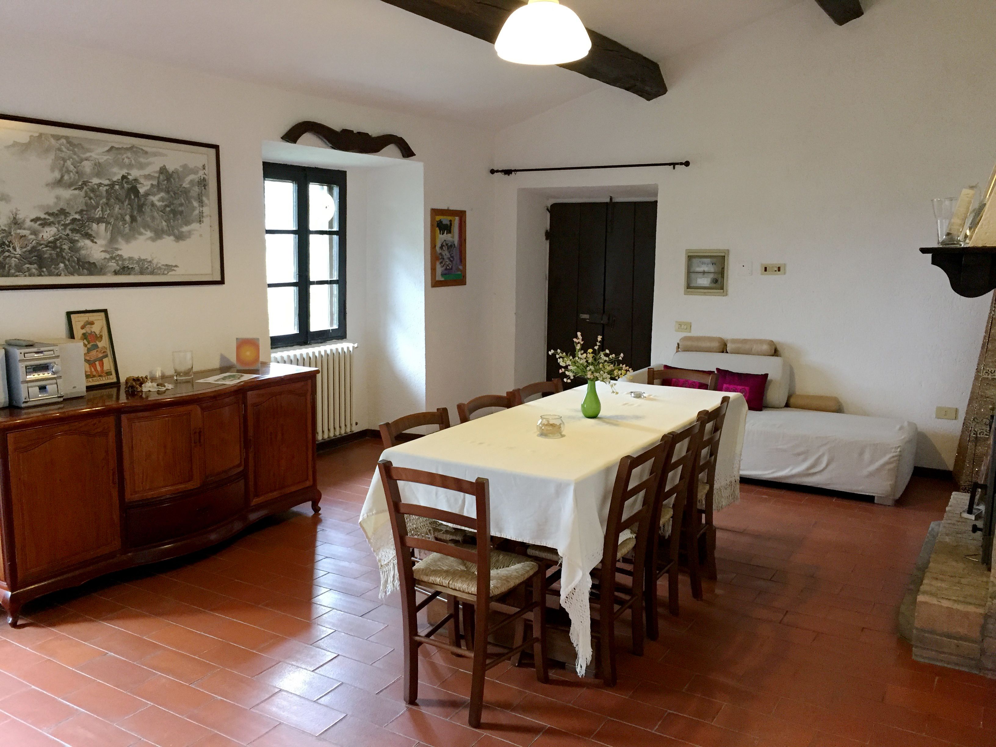 Un comedor con una mesa dispuesta para seis personas, rodeada de sillas de madera. Un aparador de madera con un radiador debajo se encuentra a la izquierda. Un cuadro cuelga en la pared sobre la ventana, y un jarrón con flores está sobre la mesa.