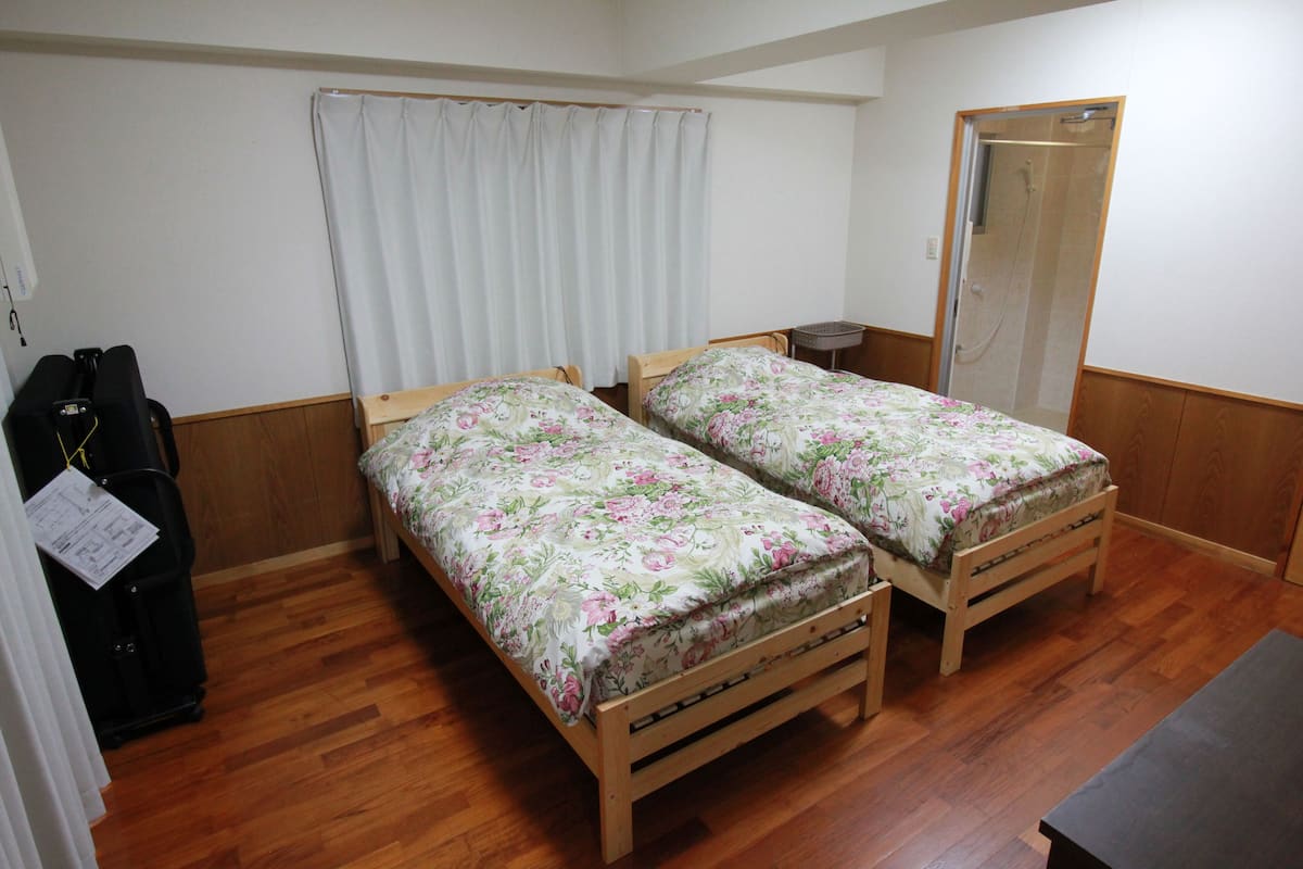 Una habitación pequeña con dos camas de madera con ropa de cama floral, un banco de madera, una mesita, una maleta y una puerta.