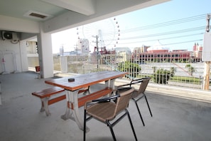 Varanda com mesa de madeira e cadeiras de vime, com vista para uma paisagem urbana com uma roda-gigante e edifícios.