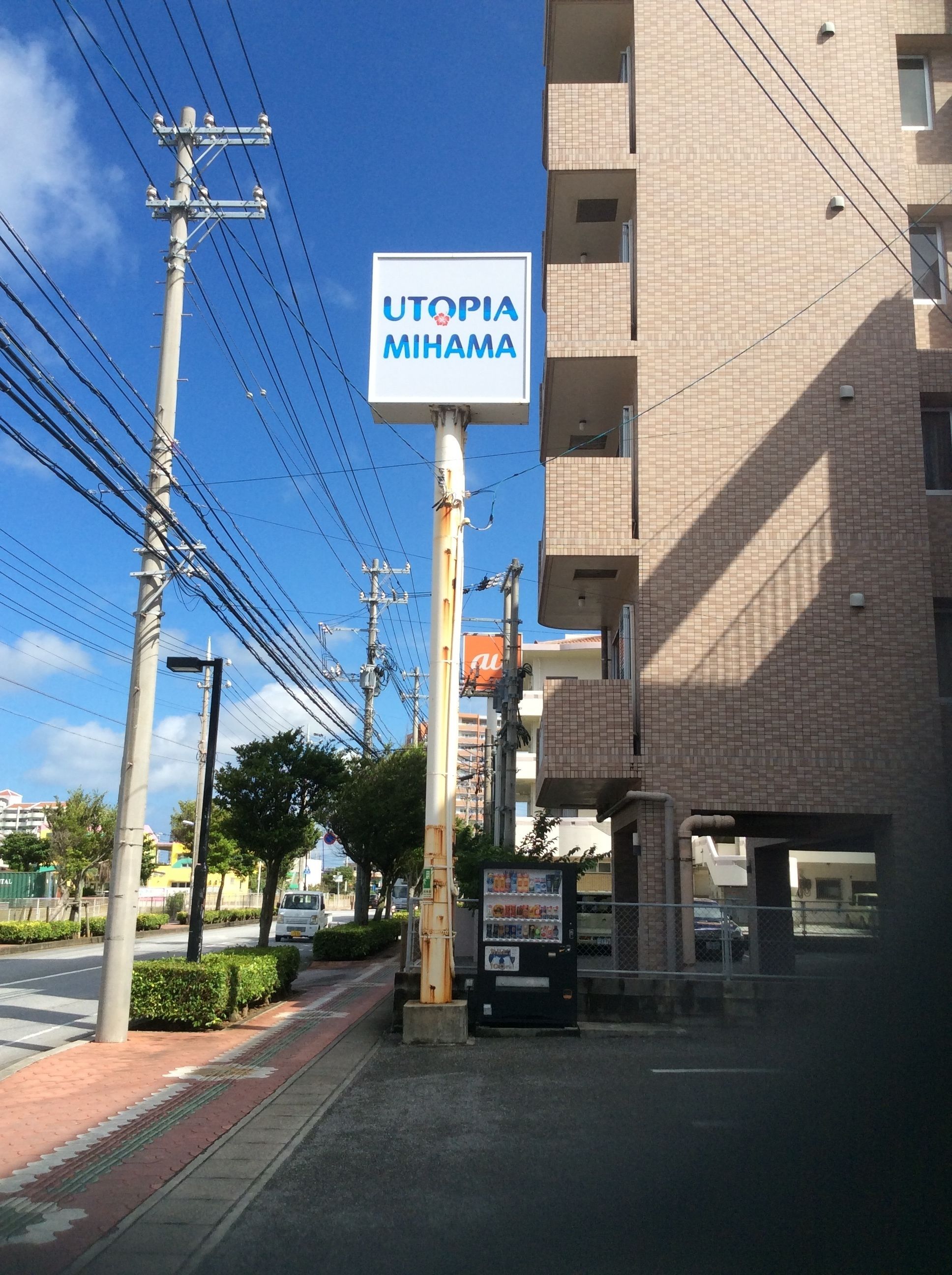 A sign for Utopia Mihama, a multi-story building with a brick facade, power lines, and a vending machine on the sidewalk.