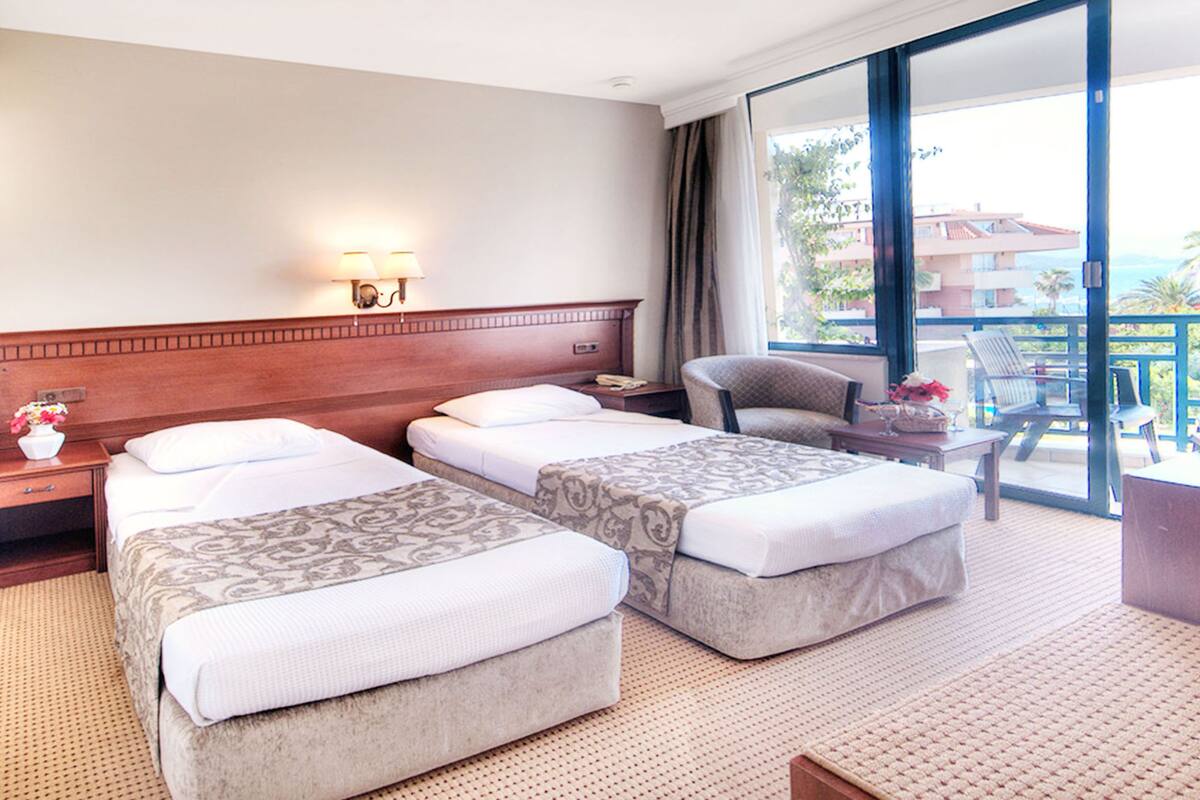 A hotel room with two beds, a sitting area with a chair and table, a large window with a view, and a nightstand with a lamp.