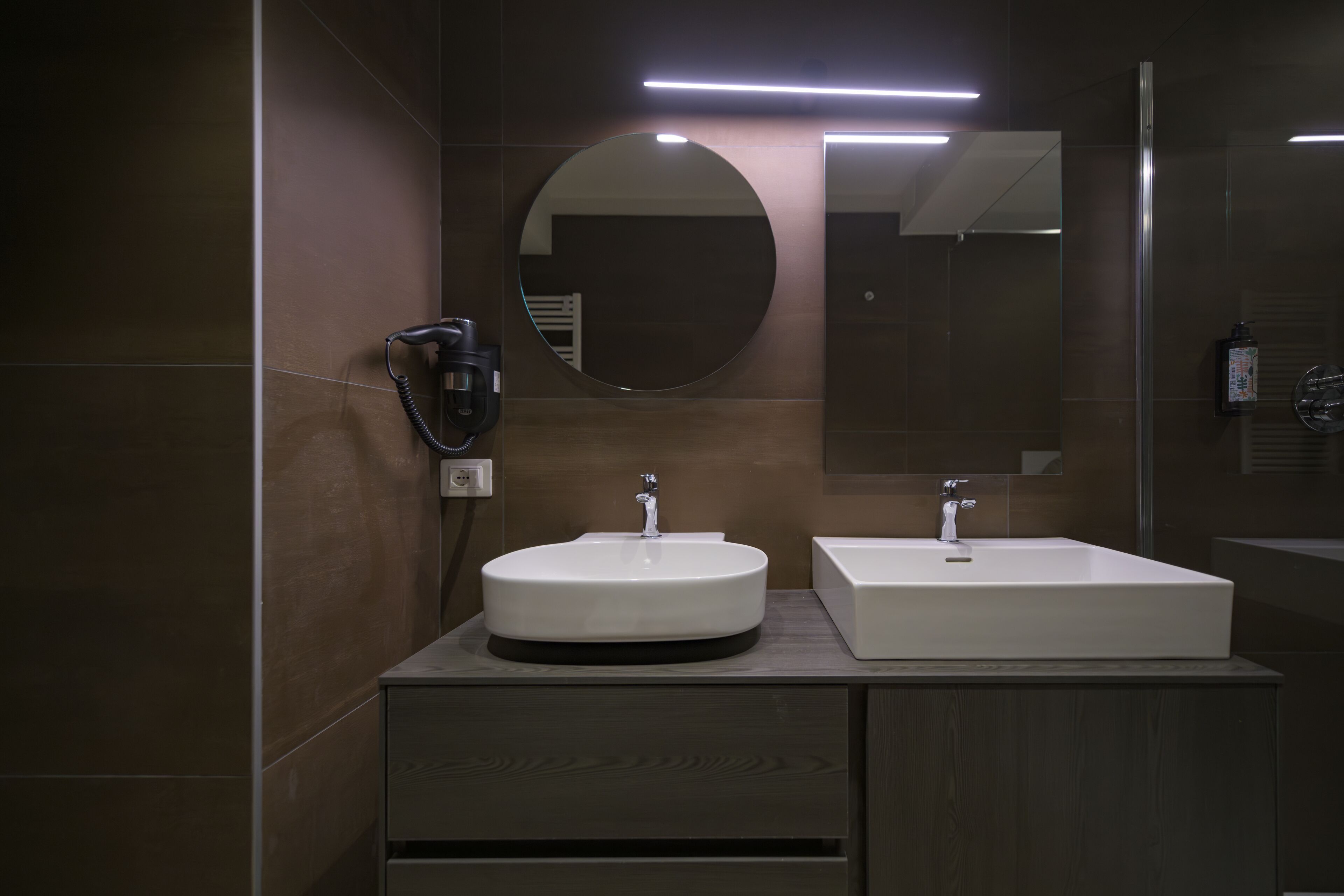 A modern bathroom with two sinks, a round mirror, and a hairdryer on the wall.