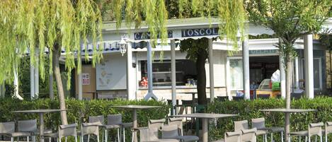Ein Straßen-Café mit Metalltischen und -stühhlen, einem überdachten Sitzbereich und einem Gebäude mit dem Schild "CAMING TOSCOLD".