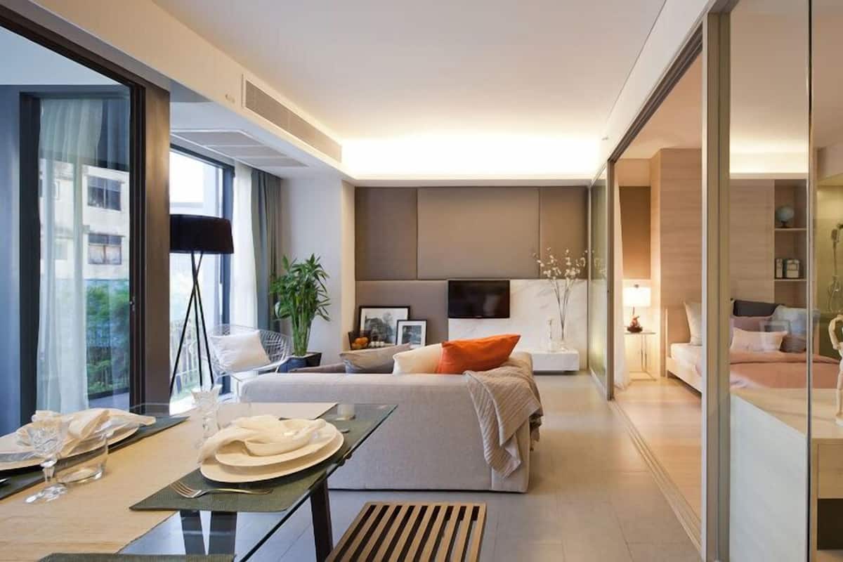 A modern hotel room with a large bed, a dining area with a table set for two, a seating area with a sofa, and a glass partition separating the living space from the bathroom.