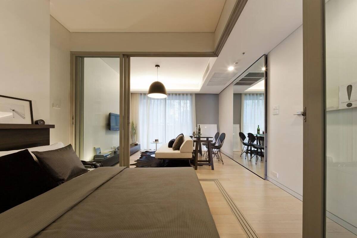 A modern hotel room with a large bed, a dining area with a table and chairs, and a glass partition separating the two spaces.