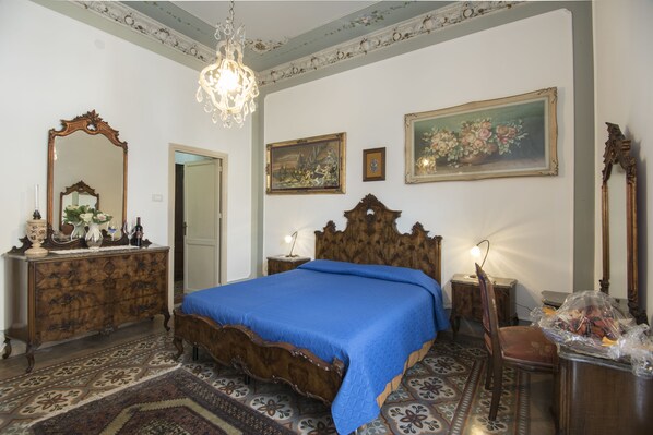 A bedroom with a large bed, ornate wooden headboard, a chandelier, a mirror, and a floral painting on the wall.