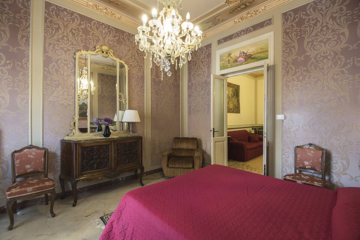 A room with a large bed, a dresser with a mirror, a chandelier, and a door leading to another room.