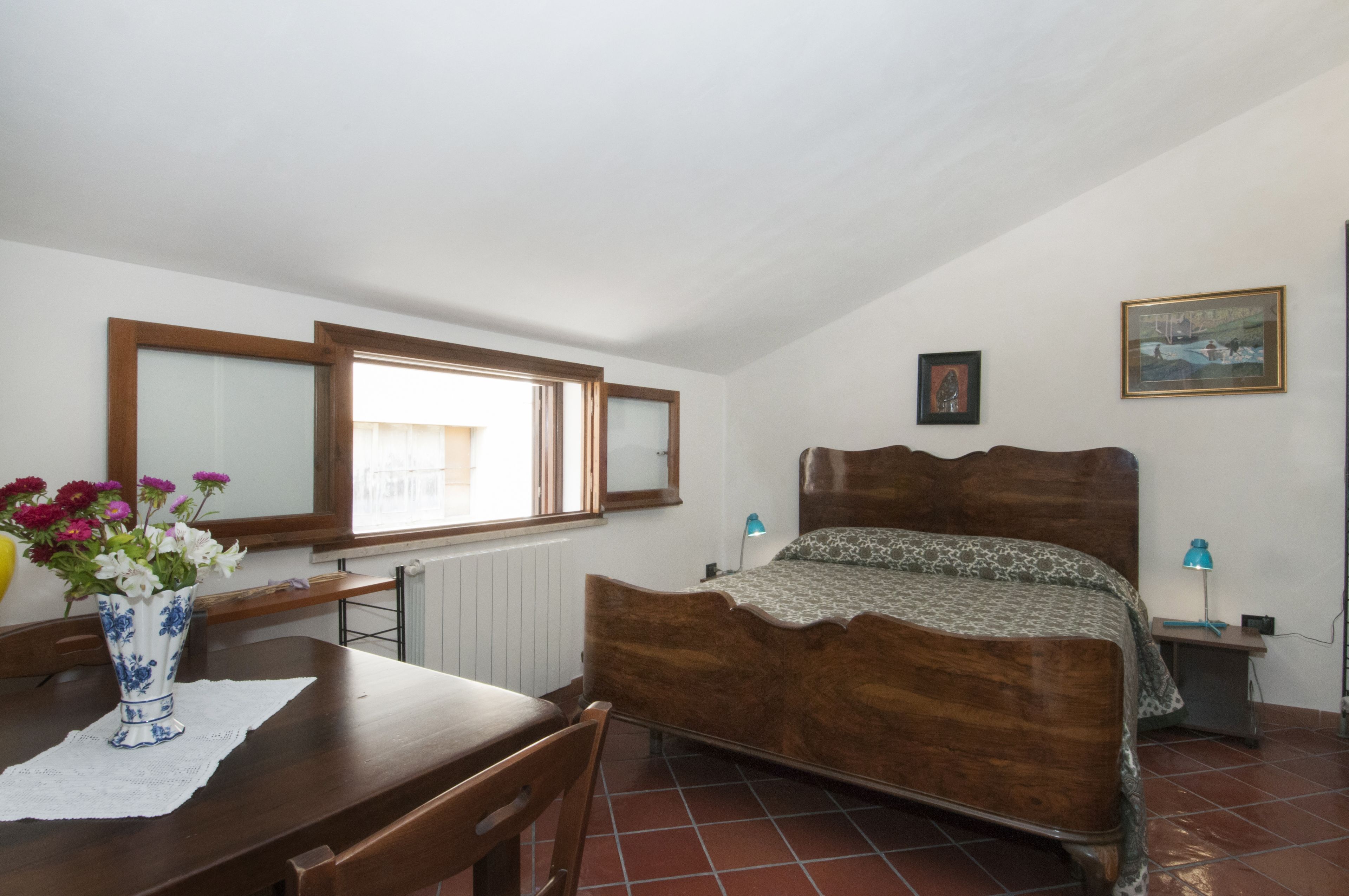 Ein Schlafzimmer mit einem hölzernen Bett, einem Fenster mit Jalousien, einem Nachttisch mit Lampe und einer Vase mit Blumen auf einem Tisch.