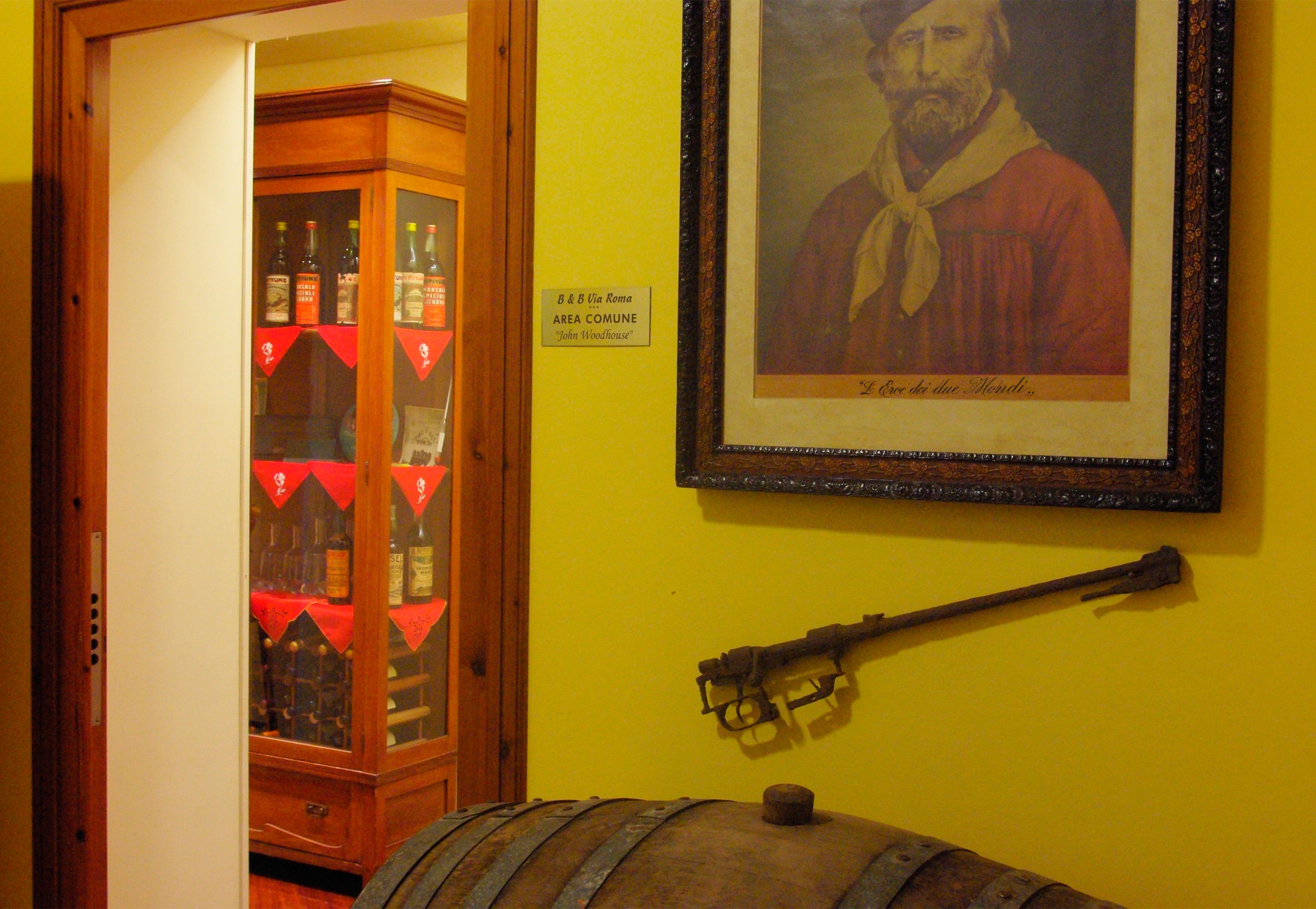 A room with a wooden cabinet displaying bottles, a framed portrait on the wall, and a mounted rifle.