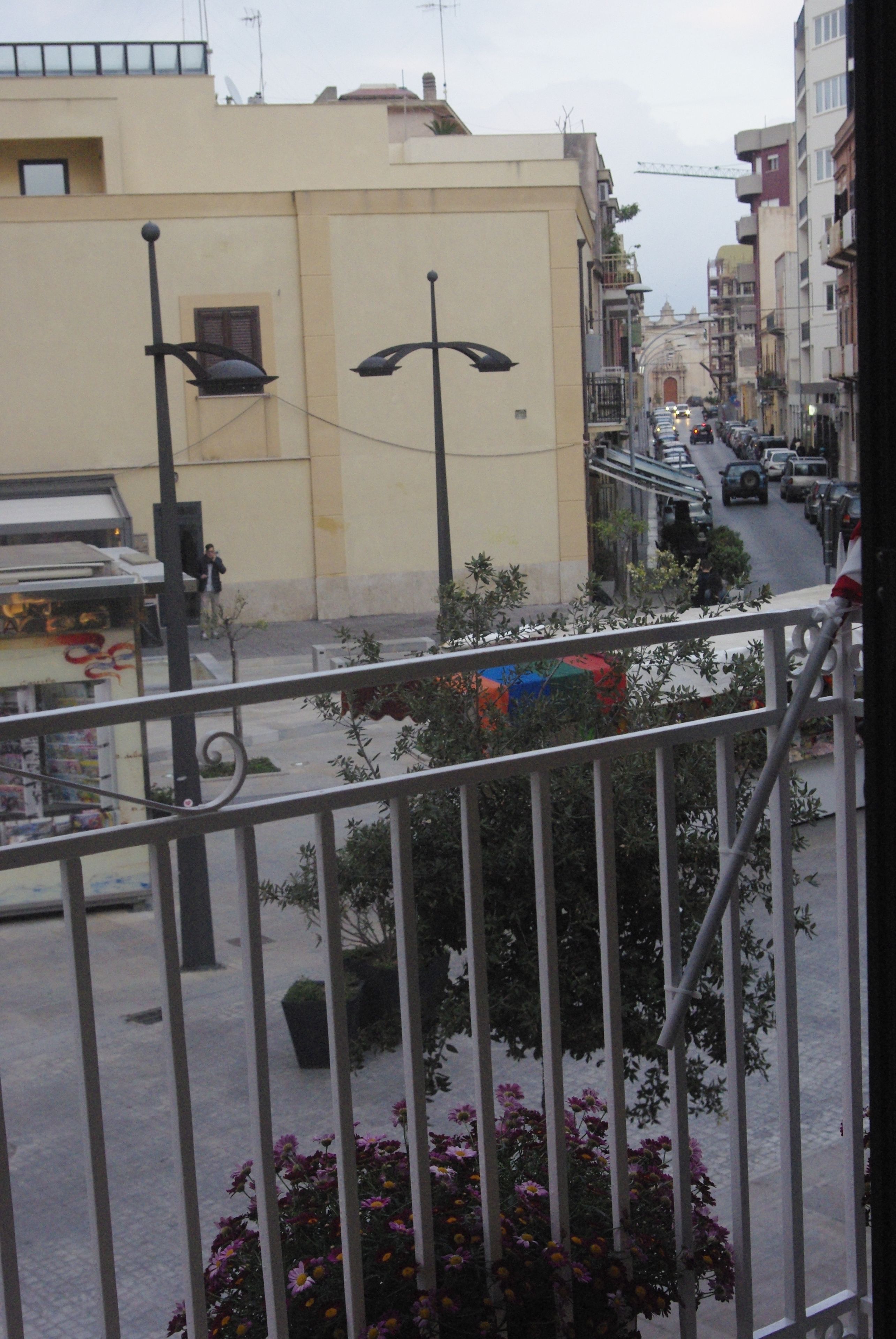 Un balcon avec des balustrades en métal donnant sur une rue où sont garées des voitures et où se trouvent des immeubles.