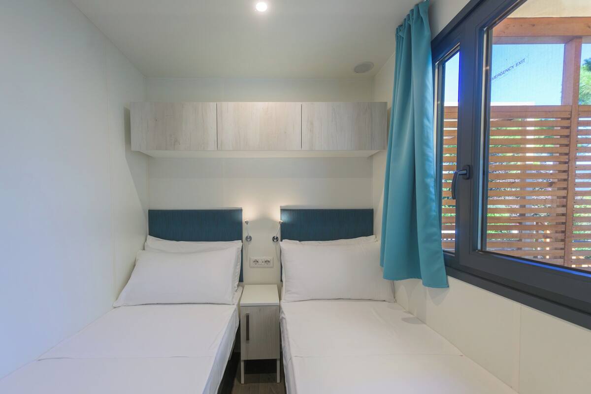 A small, modern room with two beds, a built-in wardrobe, and a window with a view of a wooden fence.