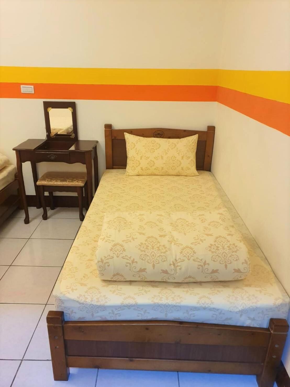 A single bed with a wooden frame and a patterned mattress, a wooden nightstand with a mirror, and a chair with a cushion.
