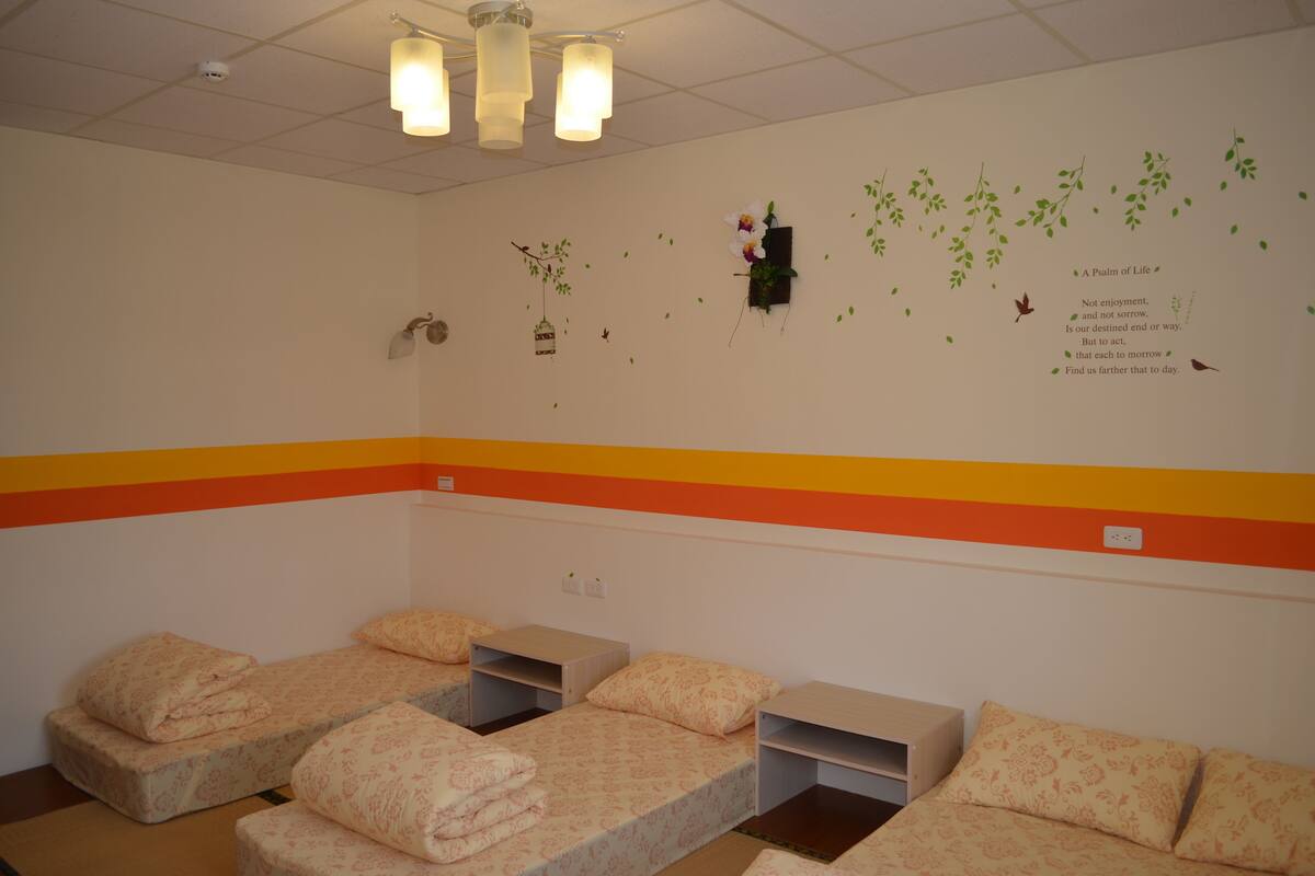 Een kamer met vier bedden, elk met een matras en kussen. De wanddecoraties omvatten een hangend horloge en een decoratief object met tekst.