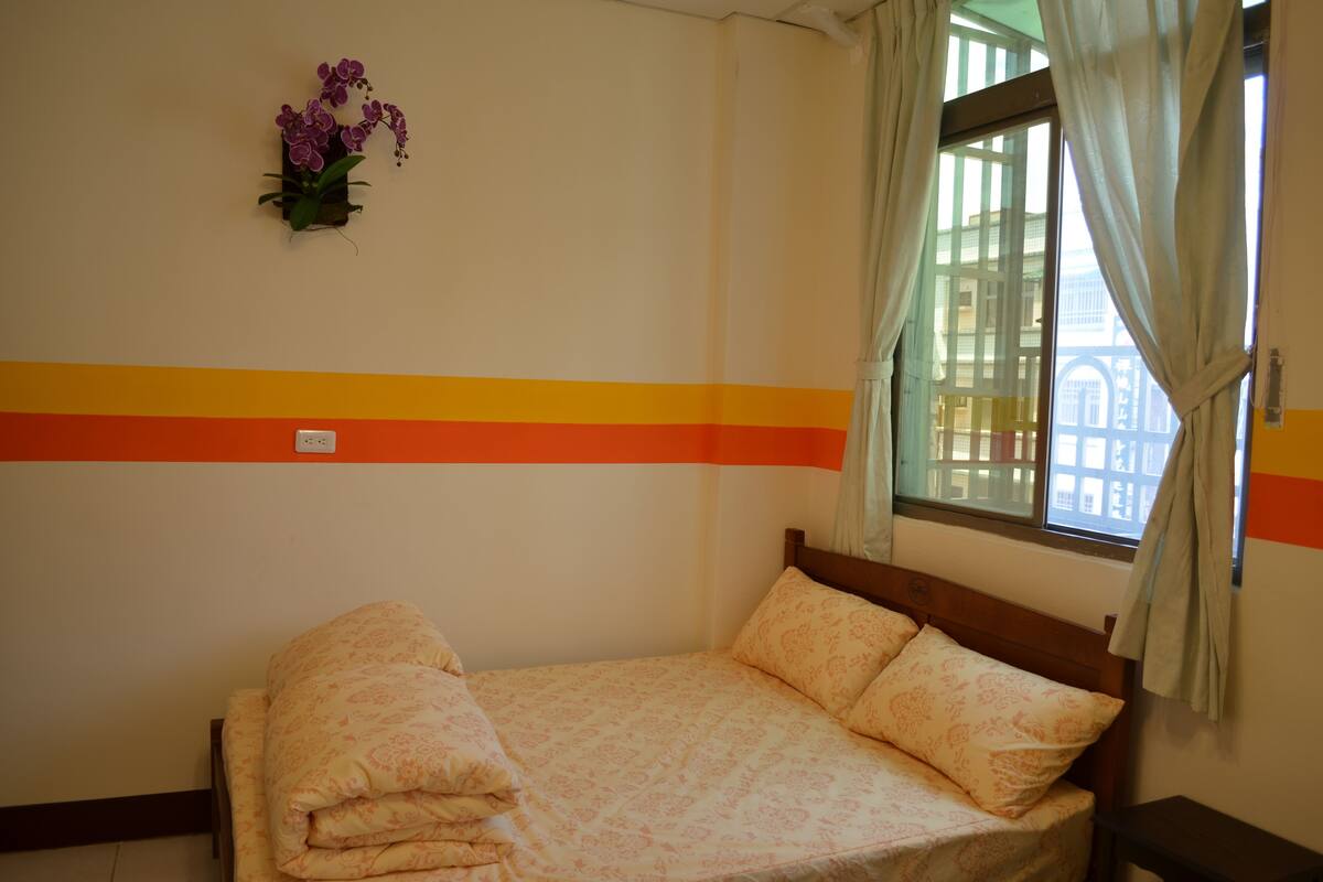 Een net opgemaakt bed met een bloemenbedekking, een aan de muur bevestigde orchidee en een raam met gordijnen.