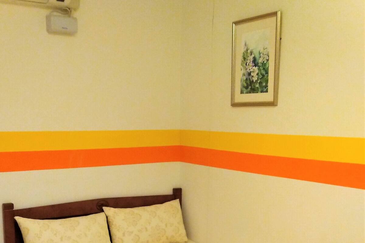 Een enkel bed met houten frame, een effen muur met gele streep, en een klein fotolijstje aan de muur.
