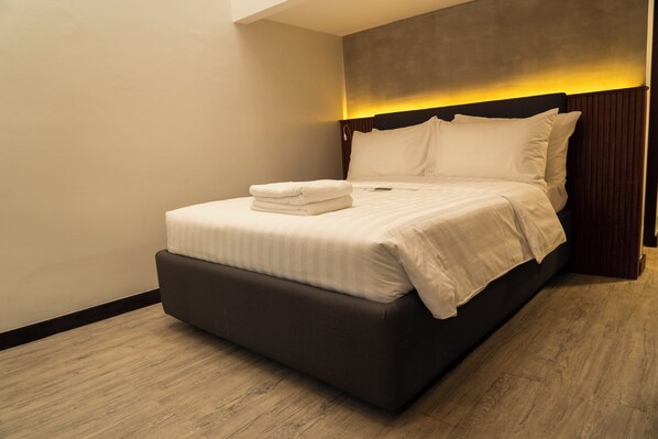 A neatly made bed with white linens and a folded towel, a dark brown headboard, and a wooden floor.