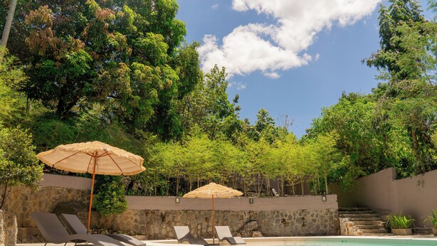 Un área de piscina con sillones de descanso y sombrillas, rodeada de muros de piedra y abundante vegetación.